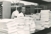 Two men between the pigeon holes and a large pile of papers for the conference. 
(IAEA Archives/Credit: Photo Ellinger)